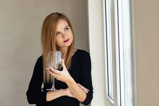 The girl stands thoughtfully at the window with a glass of red wine
