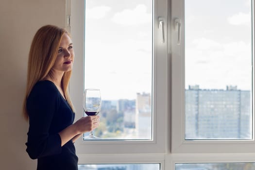 Beautiful brooding girl with a glass of red wine stands near the window
