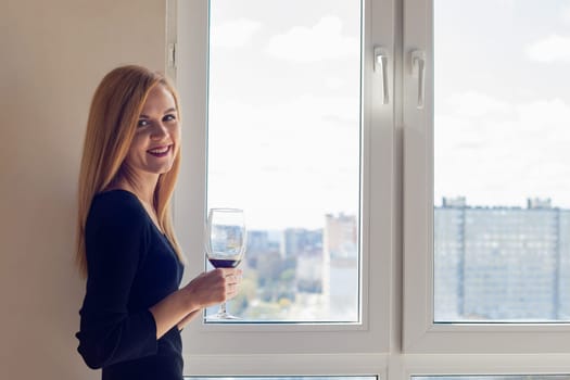 A girl stands near a window with a glass of wine