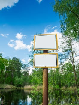 Wide-angle landscape view of a small pond framed by young trees with two signs mounted on a rusty pillar with copy space.