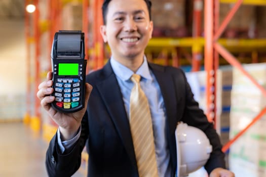 Portrait of businessman hold credit card reader in large factory and distribution warehouse environment. Business deal merger acquisitions and takeover.