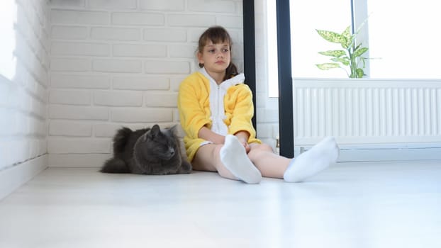 An upset girl in a yellow bathrobe sits in the corner of the room, a dark gray domestic cat sits nearby