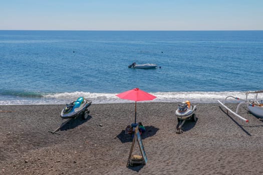 Jetski Seadoo parked on the beach. Black volcanic sand beach. In the background of the sea and moored boat. Red parasol.