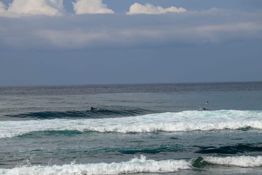 Surfers are waiting for a convenient wave on their surfboards. Tropical surfers paradise with Nyang Nyang beach on Bali island. Azure blue sky, sunny day. Wild waves are approaching the shore in rows.