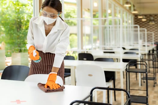 Portrait attractive asian waitress wear face mask and face shield cleaning table with alcohol and wet wipe before welcome customer. New normal hygiene restaurant concept.