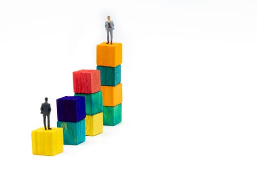 Miniature figurine posed as businessman sitting alone on a wooden cube looking up to leader on colourful stacks as business chart with focus on background, minimalist abstract concept image
