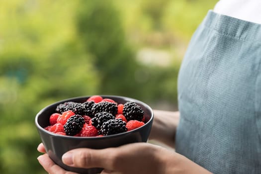 Female hands holding a bowl full of blackberries and raspberries. Unfocused background of trees.