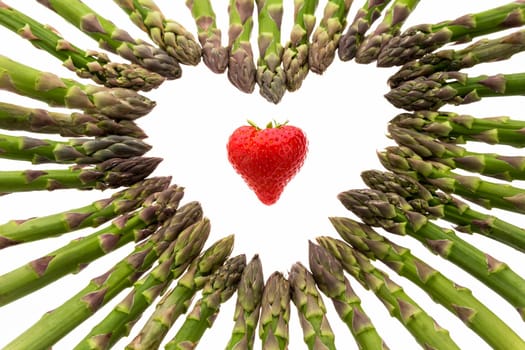 Heart-shaped red strawberry in the center of a heart made out of green asparagus tips. Color contrast between red berry and green vegetable. Close up. Top view. Cut out isolated on white background.