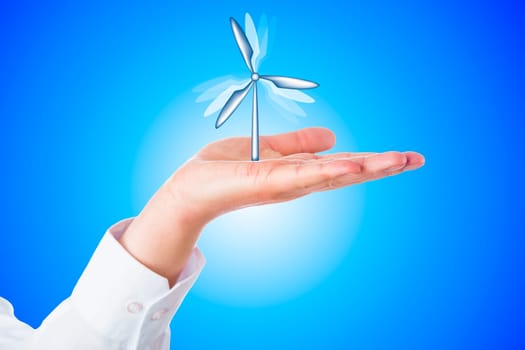Vertical wind turbine turning its rotor blades while firmly moored in the open palm of a horizontal hand. Close up over sky blue background with bright gradient center. Wind energy business concept.