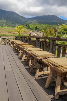 Row of wooden stools with cushions on terrace with floor made of planed wooden planks. It bumps from roughly worked beams. Background with mountains whose peaks are shrouded in thick clouds. Sunny day.