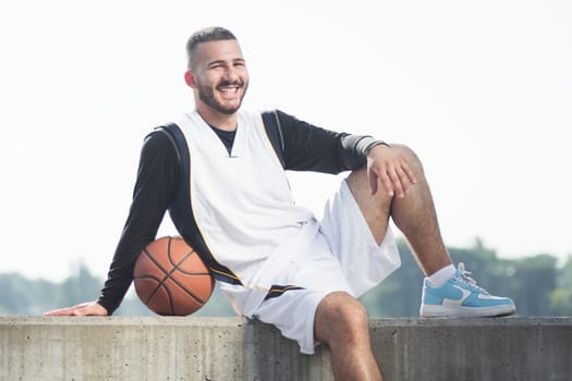 The smiling handsome young basketball player in sportswear with basketball ball is posing outdoors on white background.