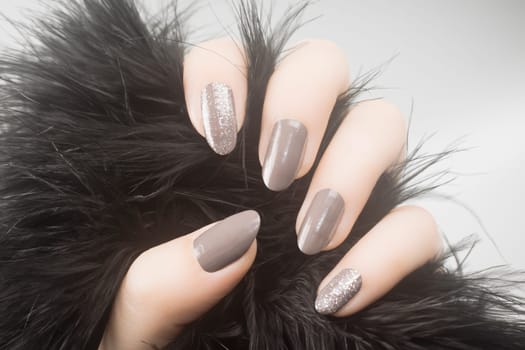 Female hand with gray nails and glittered nail polish is holding black fur on gray background, manicure concept.