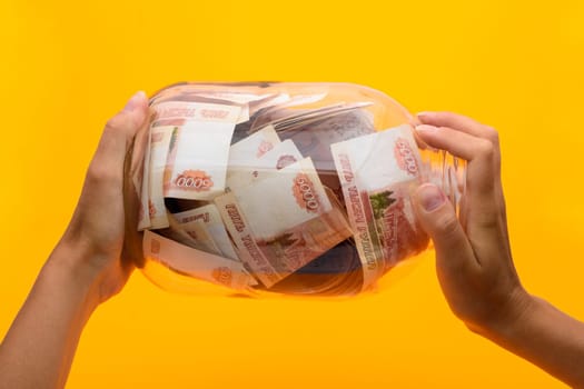 Hands holding a three-liter jar in which lie five thousandth bills, yellow background
