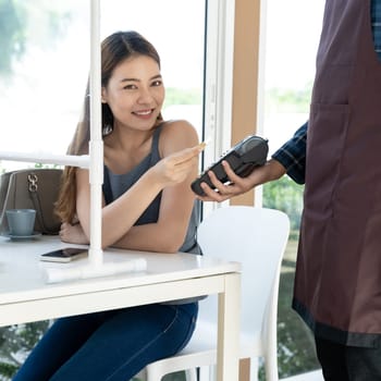 Asian woman customer hold contactless credit card for making online payment after eating out in new normal social distance restaurant. Online contactless and technology concept.