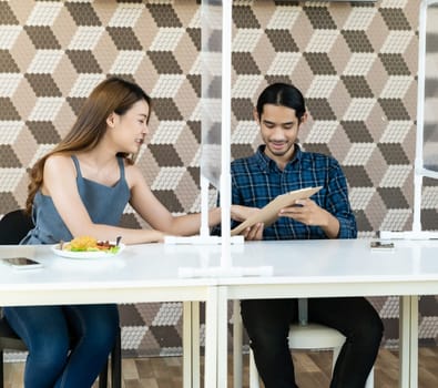 Asian young couple eating out together at new normal social distance restaurant with table shield partition reduce infection of coronavirus covid-19 pandemic. Restaurant new normal lifestyle.