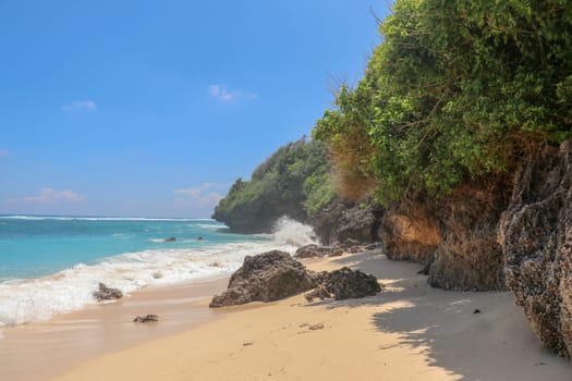 Pantai Gunung Payung beach with fine golden sand, Bali. Turquoise blue water turns into sea foam as the waves break against rocky cliffs of the coast. Sunny day with blue sky. Tourist natural concept.