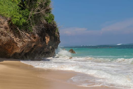 Pantai Gunung Payung beach with fine golden sand, Bali. Turquoise blue water turns into sea foam as the waves break against rocky cliffs of the coast. Sunny day with blue sky. Tourist natural concept.