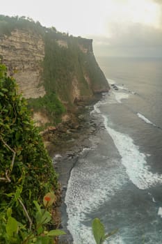 Overview panorama ocean shore, cliff. Sunset. Bali. Overwhelmed scene the flower and green-capped vertical cliff over the Indian ocean. South of Bali island, Pura Luhur Uluwatu Temple. Indonesia.