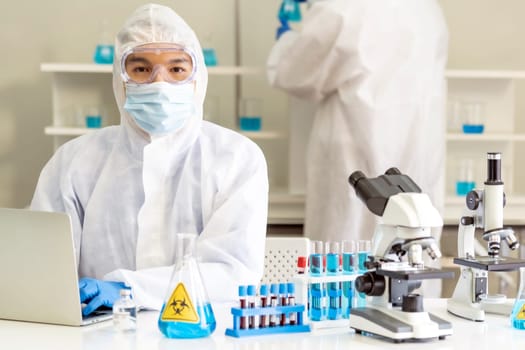 Scientists working in lab using laptop to research and develop vaccine for coronavirus covid-19 pandemic with his colleague in background. Medical Science technology and healthcare concept.