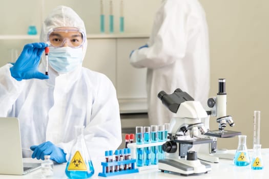 Scientists hold blood sample test tube and examine for his research and develop vaccine for coronavirus covid-19 pandemic with his colleague in background. Medical Science and technology concept.