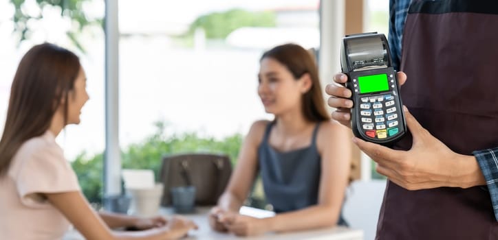 Panoramic close up of waiter hand hold credit card reader for contactless payment with customers in restaurant background. New normal restaurant contactless payment concept.