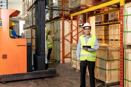 Portrait of Asian warehouse manager hold digital tablet with warehouse worker operate forklift to check inventory in background. Reopening business warehouse technology and logistic concept.