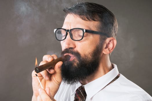 The elegant bearded man with eyeglasses is lighting a cigar on gray background.