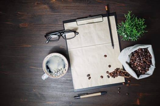 Coffee and kraft stationery. Clipboard with blank vintage letterhead, coffee cup, coffee beans, glasses, pencil and plant on wood table background. Flat lay.