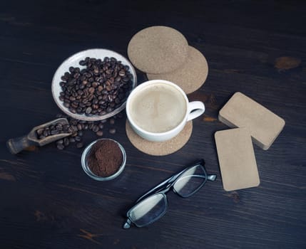 Coffee cup, roasted coffee beans, ground powder, beer coasters, blank kraft business cards and glasses on kitchen table background.