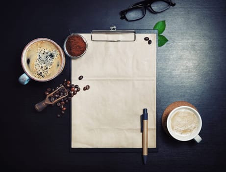 Clipboard with blank kraft letterhead, coffee cups, roasted coffee beans, pen and coffee ground on black table background. Space for text. Top view. Flat lay.