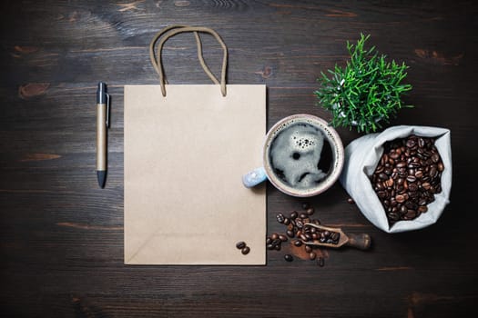 Blank kraft paper bag, coffee cup, plant, coffee beans in canvas bag and pen on wooden background. Top view. Flat lay.