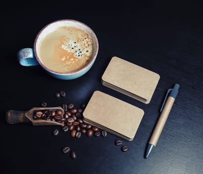 Blank kraft business cards, coffee cup, pen and coffee beans on black wooden background. Template for graphic designers portfolios.