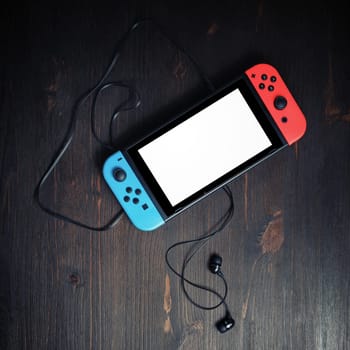 Minsk, Belarus - May 07, 2020: Nintendo Switch game console with blank white screen and headphones on vintage wooden background. Flat lay.