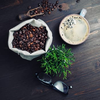 Vintage still life with coffee cup, roasted coffee beans, plant and glasses on wooden background. Flat lay.