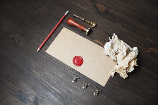 Blank kraft paper envelope with red wax seal, stamp, spoon, pencil and crumpled paper on wooden background. Mockup for your design.