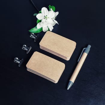 Blank kraft paper business cards, pen and flowers on black wooden background. ID template. Mock-up for branding identity for designers.