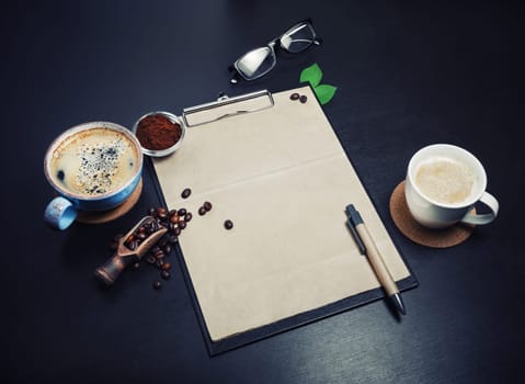 Blank retro stationery set and coffee on black table background. Clipboard with blank kraft letterhead, coffee cups, coffee beans, pen, glasses and ground powder.