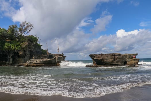 Waves shatter on a cliff at the top of which is the Hindu temple of Tanah Lot. Temple built on a rock in the sea off the coast of Bali island, Indonesia. Lush tropical vegetation on top of a cliff.