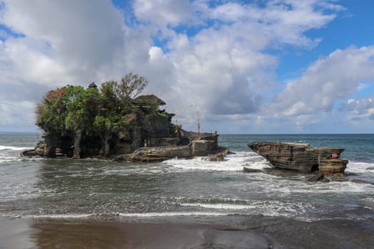 Waves shatter on a cliff at the top of which is the Hindu temple of Tanah Lot. Temple built on a rock in the sea off the coast of Bali island, Indonesia. Lush tropical vegetation on top of a cliff.