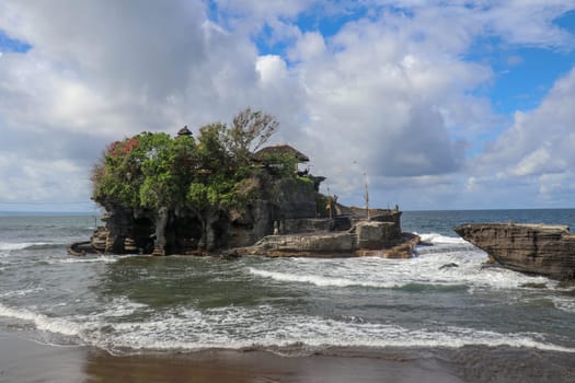 Waves shatter on a cliff at the top of which is the Hindu temple of Tanah Lot. Temple built on a rock in the sea off the coast of Bali island, Indonesia. Lush tropical vegetation on top of a cliff.