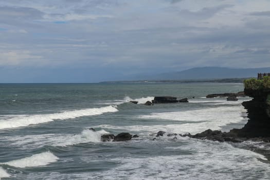 Rock cliffs in the Indian Ocean off the coast of Bali island, near Batu Bolong and tourism attraction spot Tanah Lot temple. Waves are shattering on rocks of solidified black volcanic lava. Indonesia.