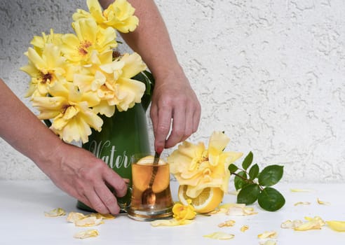woman's hand stirs lemon tea in a glass, bouquet of yellow roses stands in a green vase next to a glass of tea with lemon, scattered rose petals, creating a harmonious still life on a white table,