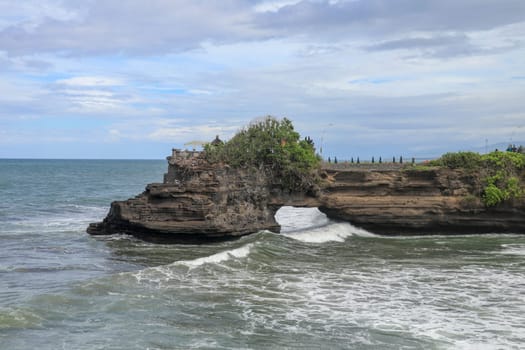 Waves arrive at a rock cliff below Batu Bolong Temple. Tanah Lot temple. Huge hole in rock cliff below Pura Batu Bolong temple. Hindu Temple on the Beatiful Rock in the morning light, Bali, Indonesia.