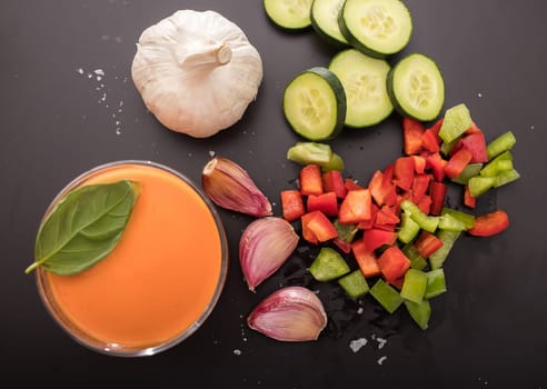 Gazpacho in two glass bowls, along with its ingredients, tomato, garlic, cucumber, red pepper and green pepper.. Top view.
