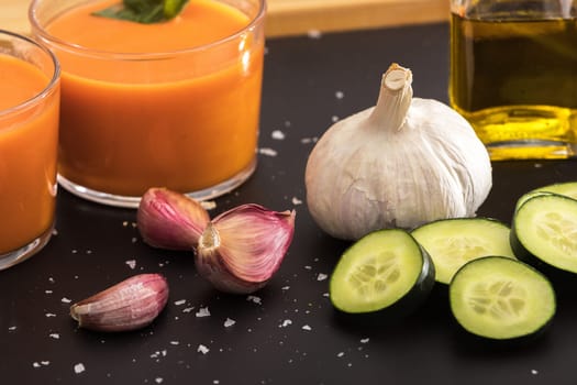 Gazpacho in two glass bowls, along with its ingredients, tomato, oil, garlic, cucumber, red pepper and green pepper.