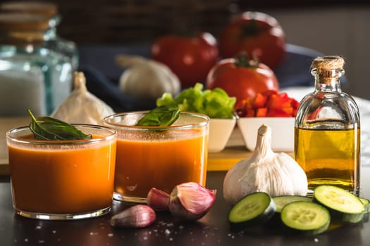 Gazpacho in two glass bowls, along with its ingredients, tomato, garlic, oil, cucumber, red pepper and green pepper.
