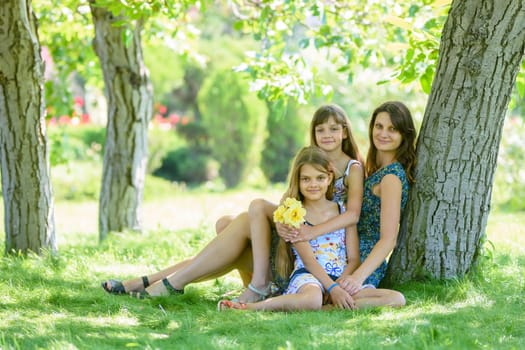 Mom with two daughters sits on green grass near tree in park