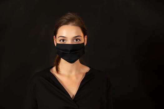 Caucasian girl in black clothes and face protection facing the camera in front of a black backdrop. Studio portrait of a young woman on dark background with copyspace for text.