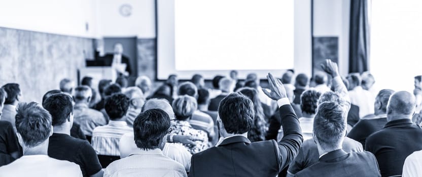 I have a question. Group of business people sitting at the chairs in conference hall. Businessman raising his arm. Conference and Presentation. Business and Entrepreneurship. Blue toned.