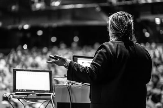 Female speaker giving a talk on corporate business conference. Unrecognizable people in audience at conference hall. Business and Entrepreneurship event. Black and white image.
